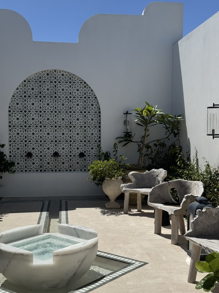 A white-walled spa courtyard with a marble fountain, tiled arch and weathered wood chairs
