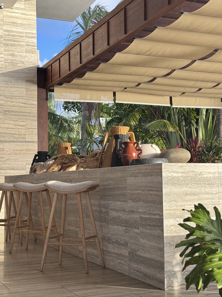 A travertine bar with ceramic vases, woven baskets and wood stools under a pergola